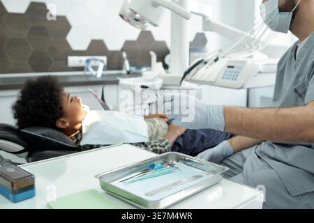 Schwarzer Junge, der mit kaukasischem männlichen Zahnarzt behandelt wird, während er auf einer Zahnarztklinik sitzt Stockfoto