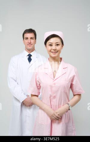 Lächelnde chinesische Krankenschwester, die mit einem kaukasischen Arzt vor grauem Hintergrund im Atelier steht Stockfoto