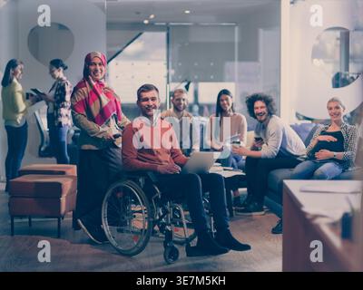 Lächelnde Geschäftsfrau aus dem Nahen Osten im Hijab stehend und helfend behinderter männlicher Kollege, der im Rollstuhl mit Laptop sitzt... Stockfoto
