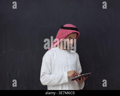 Ernsthafter Mann aus dem Nahen Osten in traditioneller arabischer Kleidung mit Tablet, während er vor schwarzem Hintergrund im Studio steht Stockfoto