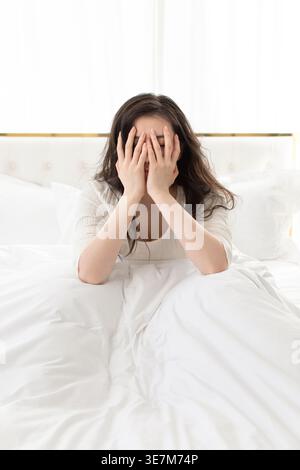 Depressive Chinesin, die das Gesicht mit den Händen bedeckt, während sie zu Hause auf dem Bett sitzt Stockfoto