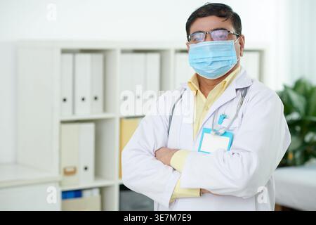 Indischer männlicher Arzt mit medizinischer Maske und Brille, der mit Armen in der Nähe des Regals im Krankenhausbüro steht Stockfoto