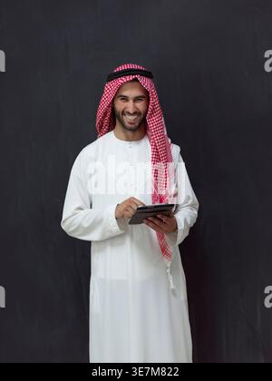 Lächelnder Mann aus dem Nahen Osten in traditioneller arabischer Kleidung mit Tablet, während er im Studio vor schwarzem Hintergrund steht Stockfoto