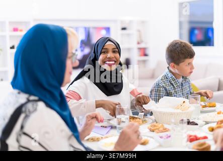 Lächelnde schwarze Frau im Hijab mit kaukasischen Freunden, die zusammen essen und zu Hause am Tisch sitzen Stockfoto