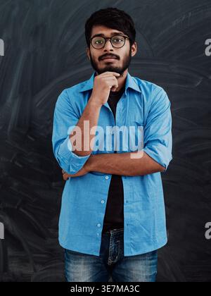 Ernster indischer Mann in blauen Langarmen mit Brille, die Hand am Kinn vor der Tafel im Klassenzimmer steht Stockfoto