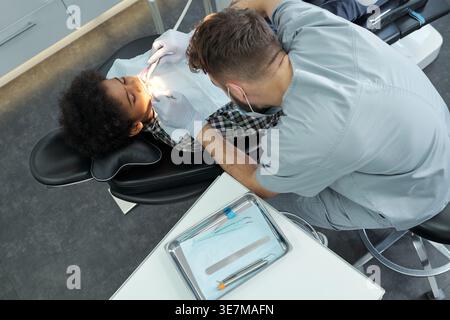 Schwarzer Junge, der mit kaukasischem männlichen Zahnarzt behandelt wird, während er auf einer Zahnarztklinik sitzt Stockfoto