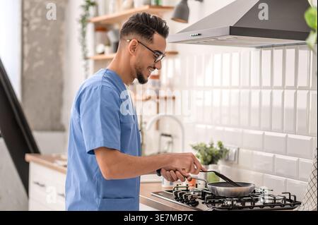 Lächelnder arabischer männlicher Pfleger in Brille kocht, während er zu Hause in der Küche steht Stockfoto