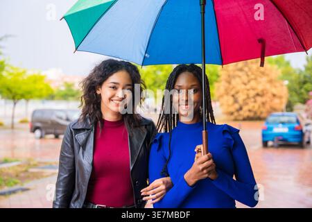 Lächelnde schwarze Frau mit Regenbogenschirm, während sie mit einer spanischen Freundin an einem regnerischen Tag auf der Straße steht Stockfoto