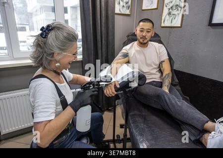 Lächelnder asiatischer Mann, der auf einem Stuhl sitzt und von einer kaukasischen Künstlerin mittleren Alters im Studio tätowiert wird Stockfoto