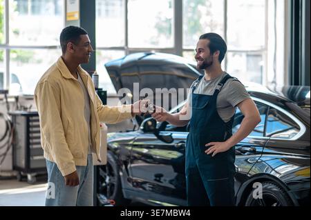 Ein afroamerikanischer Mann gibt dem männlichen kaukasier einen Autoschlüssel, der in der Autowerkstatt steht Stockfoto
