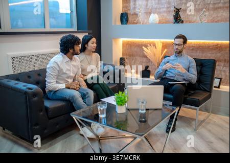 Asiatische weibliche und indische männliche Paare sitzen eng zusammen auf dem Sofa und unterhalten sich mit kaukasischen männlichen Psychotherapeuten in der Klinik Stockfoto