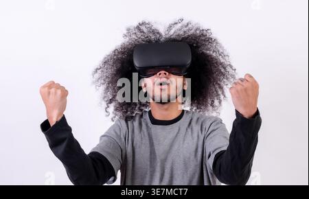 Hispanischer Mann mit lockigem Haar, der im Studio eine VR-Brille mit geballten Fäusten vor weißem Hintergrund trägt Stockfoto