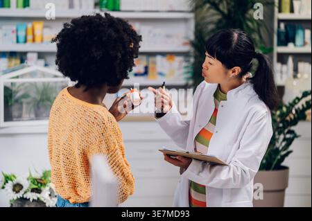Japanische Apothekerin hält Stift und Klemmbrett und spricht mit schwarzer Patientin und Kunde, die Salbenspender hält... Stockfoto