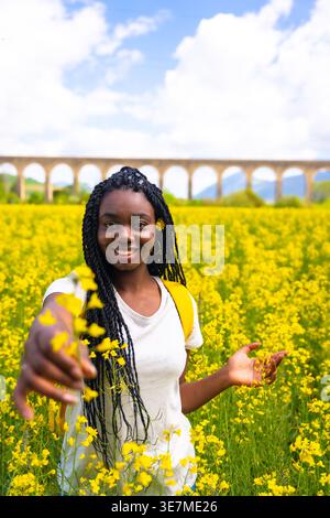 Lächelnde schwarze Frau mit Wendungen, die gelbe Blumen schenken, während sie an sonnigem Tag auf dem Feld steht Stockfoto