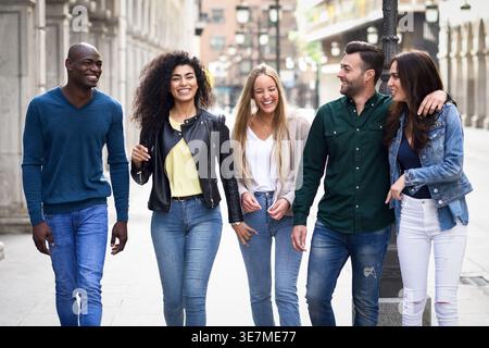 Fröhliche, vielfältige Gruppe von Freunden, die tagsüber plaudern und lachen, während sie gemeinsam auf der Stadtstraße spazieren gehen Stockfoto