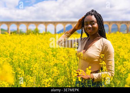 Lächelnde Schwarze Frau mit Wendungen, die den Kopf berühren, stehend auf einem Feld aus gelben Blumen an sonnigen Tagen Stockfoto