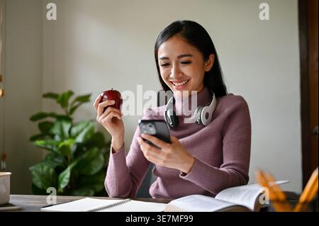 Lächelnde asiatische Frau mit Kopfhörern am Hals, die ein Smartphone mit Donut hält, während sie zu Hause mit Büchern am Tisch sitzt Stockfoto
