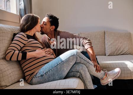 Schwarzer Mann und kaukasische schwangere Frau, die sich zu Hause auf dem Sofa küssen Stockfoto