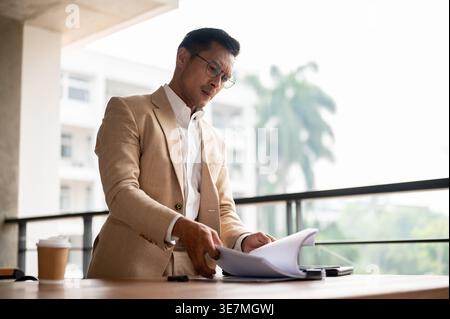 Fokussierter asiatischer Geschäftsmann mit Brille, der tagsüber in der Nähe des Geländerzauns auf dem Balkon in seiner Wohnung stand Stockfoto