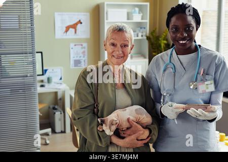 Lächelnde schwarze Tierärztin mit Klemmbrett neben der kaukasischen Seniorin, die tagsüber eine Katze in der Klinik trägt Stockfoto