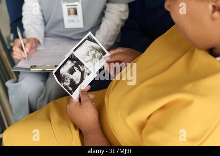 Großaufnahme einer gemischten Rasse schwangeren Frau, die sich ein Ultraschallbild ihres Babys anschaut und mit der Krankenschwester spricht, die über die medizinische Form im Wohnzimmer zu Hause schreibt Stockfoto