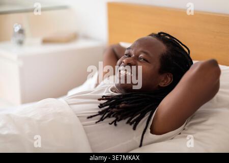 Fröhlicher Schwarzer Mann mit Dreads, der auf dem Bett liegt, mit Händen unter dem Kopf zu Hause Stockfoto