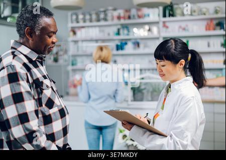 Japanische Apothekerin schreibt auf die Zwischenablage und spricht mit einem älteren schwarzen männlichen Kunden, der in der Apotheke steht Stockfoto