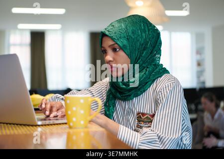 Schwarze Frau im Hijab mit Laptop, die während der Pause im Büro an der Theke mit einer Kaffeetasse sitzt Stockfoto