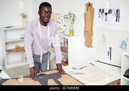 Der lächelnde schwarze Schneider trägt eine Brille und entwirft Kleidung, die am Tisch im Atelier steht Stockfoto