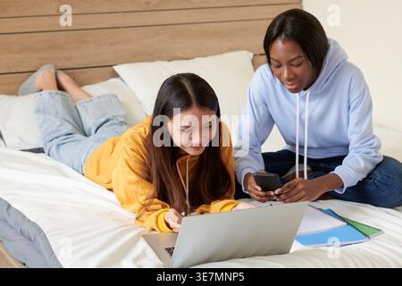 Asiatische Schülerin liegt auf ihrem Bauch und tippt auf einem Laptop mit schwarzer Klassenkameradin, die auf dem Bett sitzt und Hausaufgaben in ihrem Studentenzimmer macht Stockfoto