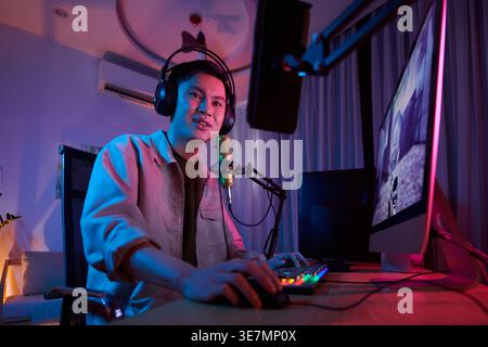 Asiatischer männlicher Online-Spiel-Streamer, der Kopfhörer trägt und Videospiele während der Live-Übertragung spielt, sitzt zu Hause auf dem Stuhl Stockfoto