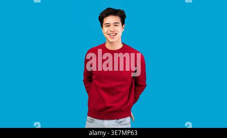 Asiatischer Mann mit rotem Pullover, der im Studio an der blauen Wand steht Stockfoto