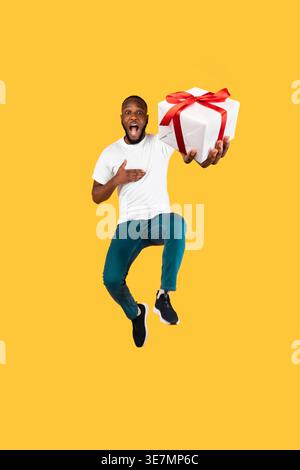 Lächelnder schwarzer Mann mit Hand auf der Brust, der Geschenk hält, während er im Studio gegen gelben Hintergrund springt Stockfoto