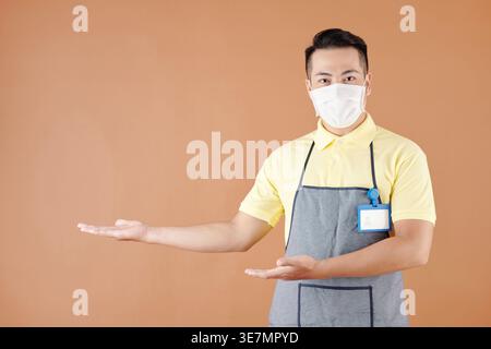 Asiatischer männlicher Café-Barista mit Schürze, der mit beiden Händen eine Willkommensgeste macht, während er im Studio vor braunem Hintergrund steht Stockfoto