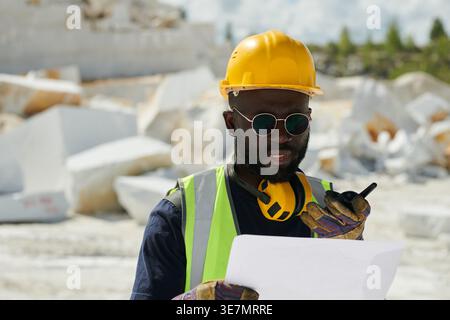 Schwarzer männlicher Ingenieur mit Schutzhelm und Sonnenbrille, der im Walkie-Talkie spricht, während er auf der Baustelle steht Stockfoto