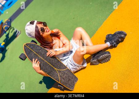 Asiatische Frau mit Hut und Sonnenbrille, die Skateboard hält und wegblickt, am sonnigen Tag auf dem Boden im Park sitzt Stockfoto