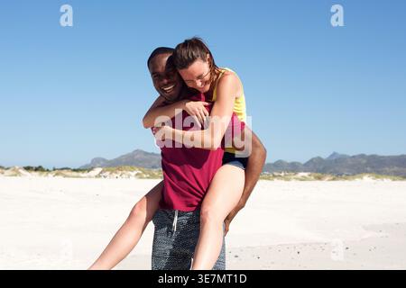 Ein lächelnder Schwarzer Mann, der an einem Sommertag am Strand einer kaukasischen Freundin einen Huckepack-Ritt gibt Stockfoto