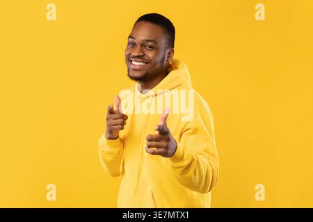 Lächelnder bärtiger Schwarzer Mann mit Hoodie, der die Finger nach vorne zeigt, während er seitlich vor gelbem Hintergrund im Studio steht Stockfoto