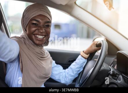 Schwarze Frau mit Hijab, die tagsüber Selfie macht, während sie im Auto sitzt Stockfoto
