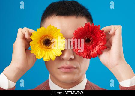 Seriöser asiatischer Mann mit rotem kariertem Anzug, der blühende rote und gelbe Blumen vor seinen Augen hält und vor blauem Hintergrund im Studio steht Stockfoto