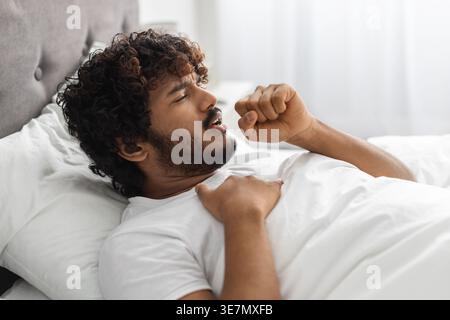 Kranker asiatischer Mann mit lockigen Haaren Husten mit Hand in Mundnähe, während er mit geschlossenen Augen auf dem Bett zu Hause lag Stockfoto