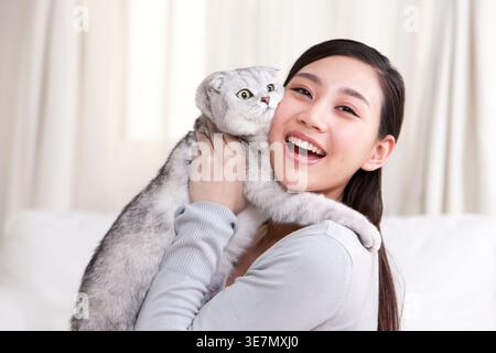 Lächelnde Chinesin, die schottische Faltkatze trägt und umarmt, während sie zu Hause auf dem Sofa sitzt Stockfoto