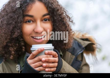 Lächelnde junge Frau mit lockigen Haaren, die warme Winterjacke trägt und im Freien Kaffee hält Stockfoto