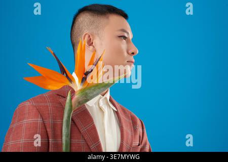 Seriöser asiatischer Mann mit Augen-Make-up und rotem Karomuster, der eine Paradiesvogel-Pflanze hält, die vor blauem Hintergrund im Studio steht Stockfoto