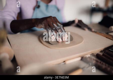 Hände des schwarzen weiblichen Lifestyleisten mit Schaber auf Ton in der Werkstatt während des Töpferkurses Stockfoto