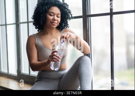 Schwarze Frau mit lockig öffnender Wasserflasche, während sie drinnen auf der Fensterbank sitzt Stockfoto