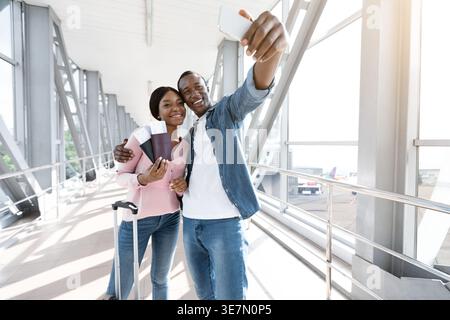 Ein lächelnder Schwarzes Paar, der Selfie macht, während er einen Reisepass mit Flugtickets auf dem Fußweg im Flughafen hält Stockfoto