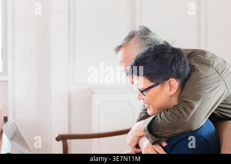 Asiatischer älterer Vater, der hinter ihm steht und einen Mann mittleren Alters umarmt, indem er zu Hause auf der Couch sitzt Stockfoto