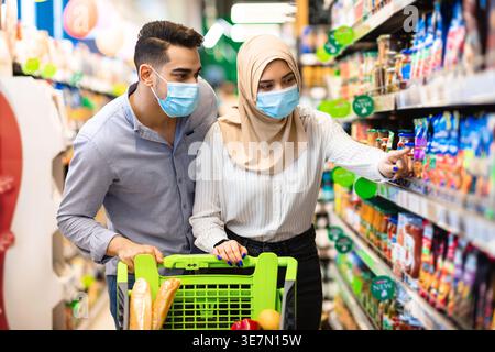Lächelndes nahöstliches Paar mit Gesichtsmaske, das Essen aus den Regalen wählt, während er mit dem Wagen im Supermarkt steht Stockfoto