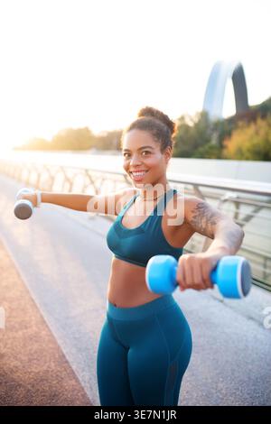Lächelnde Mixed Race Frau, die an sonnigen Tagen mit Kurzhanteln auf der Strandpromenade trainiert Stockfoto